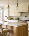 A cozy traditional kitchen featuring creamy off-white shaker cabinets, a large oak island with a honed marble top, brushed brass hardware, and a bridge faucet, illuminated by layered lighting. — view 2
