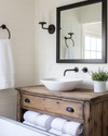 A bright farmhouse bathroom featuring a repurposed distressed wood dresser vanity with a white vessel sink, flanked by matte black barn-style sconces, above which hangs a large mirror with a simple black metal frame. A galvanized metal tub holds rolled white towels. — view 3