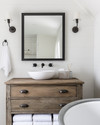 A bright farmhouse bathroom featuring a repurposed distressed wood dresser vanity with a white vessel sink, flanked by matte black barn-style sconces, above which hangs a large mirror with a simple black metal frame. A galvanized metal tub holds rolled white towels. — view 2