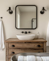 A bright farmhouse bathroom featuring a repurposed distressed wood dresser vanity with a white vessel sink, flanked by matte black barn-style sconces, above which hangs a large mirror with a simple black metal frame. A galvanized metal tub holds rolled white towels.