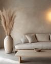A minimalist Japandi living room featuring a low-profile sofa in natural linen, a light ash wood coffee table, and a large, unglazed ceramic vase with dried pampas grass. The walls are painted in a warm grey, complemented by soft, diffused light from a paper-shade floor lamp. — view 3