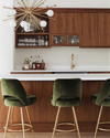 A sleek Mid-Century Modern home bar featuring warm walnut cabinetry, a pristine white quartz countertop, and polished brass bar stools with olive green velvet upholstery. A small Sputnik chandelier hangs above, illuminating a curated collection of glassware and spirits on integrated floating shelves. — view 4