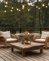 Rustic deck featuring reclaimed wood decking, a rough-hewn teak coffee table, and woven rattan armchairs with natural linen cushions. Edison bulb string lights are draped overhead, casting a warm glow, complemented by a stone fire pit in the background.