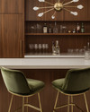 A sleek Mid-Century Modern home bar featuring warm walnut cabinetry, a pristine white quartz countertop, and polished brass bar stools with olive green velvet upholstery. A small Sputnik chandelier hangs above, illuminating a curated collection of glassware and spirits on integrated floating shelves.
