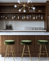A sleek Mid-Century Modern home bar featuring warm walnut cabinetry, a pristine white quartz countertop, and polished brass bar stools with olive green velvet upholstery. A small Sputnik chandelier hangs above, illuminating a curated collection of glassware and spirits on integrated floating shelves. — view 2