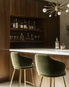 A sleek Mid-Century Modern home bar featuring warm walnut cabinetry, a pristine white quartz countertop, and polished brass bar stools with olive green velvet upholstery. A small Sputnik chandelier hangs above, illuminating a curated collection of glassware and spirits on integrated floating shelves. — view 3