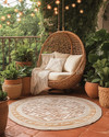 A bohemian exterior balcony featuring a terracotta floor, a woven outdoor rug with a geometric pattern, and a rattan egg chair adorned with cream cushions and a macrame throw. Lush green potted plants in various terracotta and woven planters are arranged around, with warm string lights draped overhead. — view 4