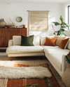 A bohemian great room blending vintage and modern elements, featuring a cream sectional sofa, a mid-century teak credenza, layered jute and sheepskin rugs, and a mix of terracotta and sage green accents.