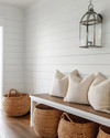 A bright coastal mudroom featuring a white shiplap wall, a light-toned reclaimed wood bench, oversized woven rattan baskets for storage, and a striking glass-encased brushed nickel lantern pendant light illuminating the space.