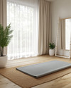 A Scandinavian home gym designed for a seasonal refresh, featuring light wood flooring, white walls, and a large window with sheer curtains. A natural jute rug is placed under a light grey yoga mat, with a potted fern in a white ceramic planter and a simple birch-framed mirror nearby. — styling and décor detail