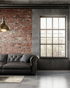 An industrial living room with a cohesive color story featuring a charcoal leather sofa, exposed brick wall, and a large metal-framed window. Warm grey walls, a polished concrete floor, and an aged brass pendant light add to the calm, urban aesthetic. — styling and décor detail
