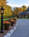 A charming farmhouse driveway leading to a home, featuring a gravel path edged with natural fieldstone, flanked by seasonal planters with colorful mums and pumpkins, and illuminated by dark bronze lantern-style lights on posts. — close-up of textures and materials
