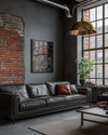 An industrial living room with a cohesive color story featuring a charcoal leather sofa, exposed brick wall, and a large metal-framed window. Warm grey walls, a polished concrete floor, and an aged brass pendant light add to the calm, urban aesthetic.