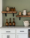 Calm farmhouse home bar featuring warm white shaker cabinets, a honed light grey soapstone countertop, and open reclaimed wood shelving. Amber glass bottles, ceramic pitchers, and matte black hardware complement the serene sage green accent wall. — styling and décor detail