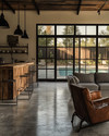 Industrial poolhouse interior featuring polished concrete floors, a reclaimed wood bar, and black steel framed windows looking out to a pool. A charcoal linen sofa, vintage leather armchairs, and matte black industrial pendant lights create a vintage-modern fusion. — close-up of textures and materials