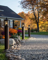 A charming farmhouse driveway leading to a home, featuring a gravel path edged with natural fieldstone, flanked by seasonal planters with colorful mums and pumpkins, and illuminated by dark bronze lantern-style lights on posts. — alternate angle