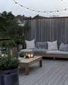 A serene Scandinavian deck with light-toned wooden flooring, a low-profile wooden lounge set with off-white cushions and a chunky knit grey wool throw, minimalist galvanized steel planters with evergreens, and soft string lights overhead. — close-up of textures and materials