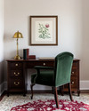 A traditional small home office featuring a dark wood secretary desk with a brass lamp, a comfortable upholstered desk chair in deep green, and a classic Persian rug in muted red and cream tones. A framed botanical print hangs on the wall. — alternate angle