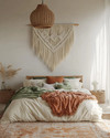 Boho bedroom with a low-slung bed, warm woven rattan pendant light, macrame wall hanging above the bed, and layered textiles in cream, terracotta, and soft green accents. — alternate angle