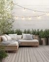 A serene Scandinavian deck with light-toned wooden flooring, a low-profile wooden lounge set with off-white cushions and a chunky knit grey wool throw, minimalist galvanized steel planters with evergreens, and soft string lights overhead.