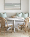 A serene coastal breakfast nook featuring a round bleached oak table with two woven rattan chairs and a light grey upholstered bench, accented with pale seafoam green throw pillows and a natural jute rug. — styling and décor detail