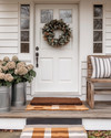 A charming farmhouse exterior with a white front door, layered buffalo check and coir doormats, and a dried eucalyptus wreath. Galvanized metal planters with hydrangeas flank the entrance, beside a distressed wooden bench with a striped throw pillow. — alternate angle