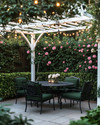 A traditional backyard patio area featuring a wrought iron dining set with dark green cushions, surrounded by lush green boxwood hedges and climbing pink roses on a white trellis, all illuminated by warm string lights overhead. — close-up of textures and materials