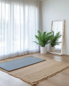 A Scandinavian home gym designed for a seasonal refresh, featuring light wood flooring, white walls, and a large window with sheer curtains. A natural jute rug is placed under a light grey yoga mat, with a potted fern in a white ceramic planter and a simple birch-framed mirror nearby.