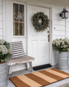 A charming farmhouse exterior with a white front door, layered buffalo check and coir doormats, and a dried eucalyptus wreath. Galvanized metal planters with hydrangeas flank the entrance, beside a distressed wooden bench with a striped throw pillow. — styling and décor detail