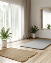 A Scandinavian home gym designed for a seasonal refresh, featuring light wood flooring, white walls, and a large window with sheer curtains. A natural jute rug is placed under a light grey yoga mat, with a potted fern in a white ceramic planter and a simple birch-framed mirror nearby. — alternate angle