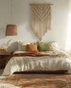 Boho bedroom with a low-slung bed, warm woven rattan pendant light, macrame wall hanging above the bed, and layered textiles in cream, terracotta, and soft green accents. — styling and décor detail