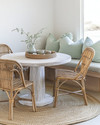 A serene coastal breakfast nook featuring a round bleached oak table with two woven rattan chairs and a light grey upholstered bench, accented with pale seafoam green throw pillows and a natural jute rug. — alternate angle