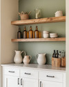 Calm farmhouse home bar featuring warm white shaker cabinets, a honed light grey soapstone countertop, and open reclaimed wood shelving. Amber glass bottles, ceramic pitchers, and matte black hardware complement the serene sage green accent wall. — alternate angle