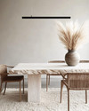 A modern dining room featuring a bleached oak dining table with a live edge, woven rattan dining chairs, and a large, textured cream area rug. A matte black linear pendant light hangs above, and a minimalist ceramic vase with dried pampas grass adorns the table. — alternate angle