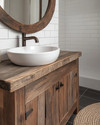 A cozy rustic bathroom featuring a reclaimed wood vanity with a white ceramic vessel sink, dark bronze faucet, and a large round mirror with a distressed wooden frame. White subway tile lines the lower wall, and a woven jute rug adds warmth to the dark grey floor tiles. — alternate angle