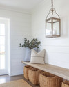 A bright coastal mudroom featuring a white shiplap wall, a light-toned reclaimed wood bench, oversized woven rattan baskets for storage, and a striking glass-encased brushed nickel lantern pendant light illuminating the space. — alternate angle