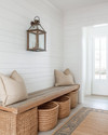 A bright coastal mudroom featuring a white shiplap wall, a light-toned reclaimed wood bench, oversized woven rattan baskets for storage, and a striking glass-encased brushed nickel lantern pendant light illuminating the space. — styling and décor detail