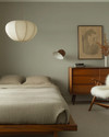 A Japandi bedroom blending vintage and modern, featuring a low oak bed, a mid-century modern teak dresser, a modern rice paper pendant light, and a vintage spindle-back chair with a sheepskin throw, all in muted tones of soft grey, beige, and sage green. — alternate angle