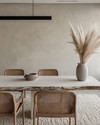 A modern dining room featuring a bleached oak dining table with a live edge, woven rattan dining chairs, and a large, textured cream area rug. A matte black linear pendant light hangs above, and a minimalist ceramic vase with dried pampas grass adorns the table. — styling and décor detail