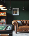 Rustic game room showcasing a blend of vintage and modern elements, with deep forest green walls, a classic leather chesterfield sofa, a vintage arcade machine, and industrial pendant lights illuminating a sleek poker table. — alternate angle
