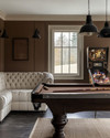 Farmhouse game room with a dark wood billiard table, a cream linen Chesterfield sofa, matte black metal lighting fixtures, and a vintage pinball machine against a wall painted in 'Swiss Coffee'. — alternate angle