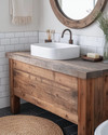 A cozy rustic bathroom featuring a reclaimed wood vanity with a white ceramic vessel sink, dark bronze faucet, and a large round mirror with a distressed wooden frame. White subway tile lines the lower wall, and a woven jute rug adds warmth to the dark grey floor tiles.