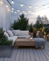 A serene Scandinavian deck with light-toned wooden flooring, a low-profile wooden lounge set with off-white cushions and a chunky knit grey wool throw, minimalist galvanized steel planters with evergreens, and soft string lights overhead. — alternate angle