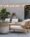 A minimalist boho deck featuring light teak wood lounge chairs with off-white cushions, subtle woven rattan accents, and large concrete planters holding a Fiddle-Leaf Fig and various succulents, all under warm string lights. — alternate angle