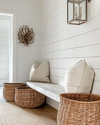 A bright coastal mudroom featuring a white shiplap wall, a light-toned reclaimed wood bench, oversized woven rattan baskets for storage, and a striking glass-encased brushed nickel lantern pendant light illuminating the space. — close-up of textures and materials