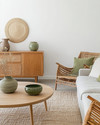 A Scandinavian Craft living room blending vintage and modern, featuring a light oak coffee table, a mid-century teak sideboard, a contemporary sofa in a light grey linen, and a woven rattan armchair, accented with handmade pottery and muted green textiles. — alternate angle