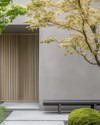 A serene Japandi front yard with a cohesive color palette, featuring a light grey exterior wall, natural blonde wood slatted screen, a minimalist dark grey bench, and subtle green Japanese maples and moss accents.
