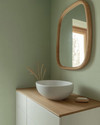 A serene Japandi powder room featuring a floating white oak vanity with a matte white vessel sink, an organically shaped mirror with a slim natural wood frame, and a soft sage green limewash wall. — styling and décor detail