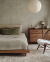 A Japandi bedroom blending vintage and modern, featuring a low oak bed, a mid-century modern teak dresser, a modern rice paper pendant light, and a vintage spindle-back chair with a sheepskin throw, all in muted tones of soft grey, beige, and sage green.