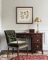 A traditional small home office featuring a dark wood secretary desk with a brass lamp, a comfortable upholstered desk chair in deep green, and a classic Persian rug in muted red and cream tones. A framed botanical print hangs on the wall. — styling and décor detail